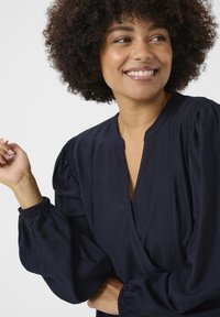 Blusa marina con un escote en V profundo, mangas largas voluminosas con puños fruncidos y una tela texturizada. Diseño simple y elegante.