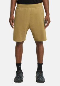 Pantaloni corti in felpa color khaki con cintura elastica, tasche laterali e una texture liscia. Abbinati a calze e scarpe nere, con un motivo mimetico.