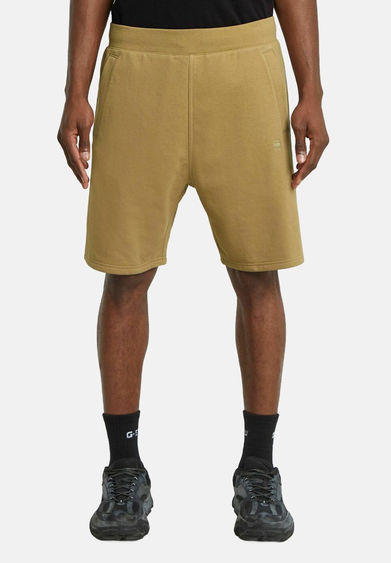 Pantaloni corti in felpa color khaki con cintura elastica, tasche laterali e una texture liscia. Abbinati a calze e scarpe nere, con un motivo mimetico.