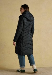 Melnā pūkaini mētelis ar kapuci, ar ševronu rakstu. Valkāts virs gaiši ziliem sarullētiem džinsiem un zaļiem sporta apaviem. Tekstūra, gluda apdare.