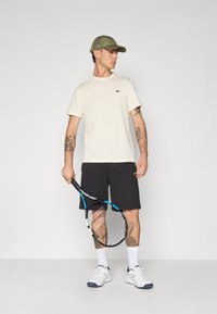 Heled beeziv t-särk väikese rohelise logo, mustad lühikesed püksid, valged sokid ja tennisereket. Viimistletud oliivrohelise kübaraga.