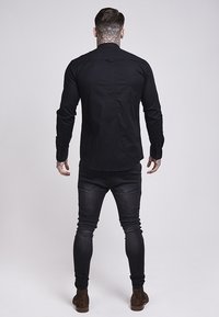 Zwart shirt met lange mouwen; nauwsluitend ontwerp met een gebogen zoom en een detail van een achternaad, gecombineerd met donkerblauwe slim-fit jeans en bruine laarzen.