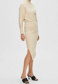 Beige sweater dress with a mock neck, ribbed texture, and side slit. Paired with black open-toe mules featuring a wide strap.