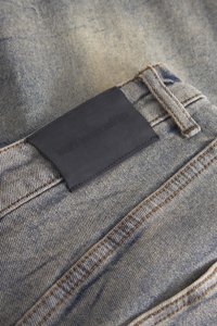 Denimjeans hebben een lichtblauwe wassing met een gestructureerd oppervlak. Een zwart leren label is aan de achterkant van de tailleband genaaid, wat zorgt voor merkindentificatie.