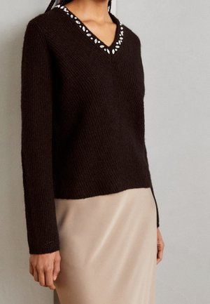 Pull noir à manches longues en V avec des ornements argentés sur le col, porté avec une jupe beige sur un fond gris uni.