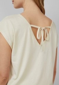 Vrouw in een crèmekleurige top met korte mouwen en een V-hals aan de achterkant die met een klein strikje vastgebonden is.