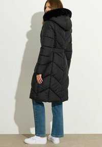 Sort puffy frakke med h�ætte, med et faux fur-krave, chevron søm og snøre i taljen. Bæres over blå jeans og hvide sneakers.