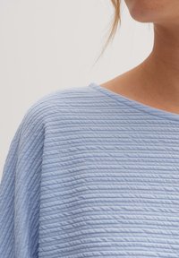 Jasnoniebieska, teksturowana bluzka z żeberkowym wzorem, szerokim dekoltem w serek oraz miękkim, zwiewnym materiałem. Zbliżenie na obszar ramienia.