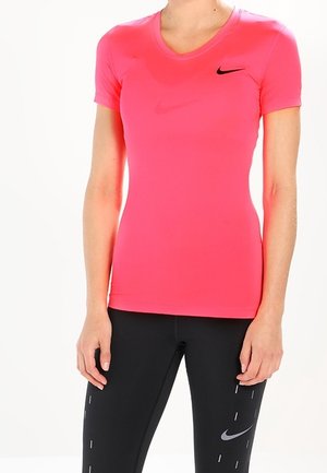 Kobieta ubrana w jasnoróżowy, krótki rękaw sportowej koszulki Nike oraz czarne legginsy Nike z odblaskowymi detalami, stojąca na jednolitym białym tle.