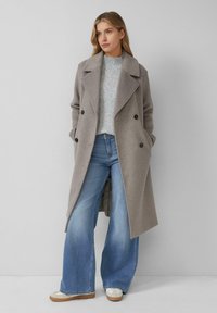 Vrouw in een grijze dubbele wolcoat, lichtgrijze gebreide sweater, wijdpeervormige blauwe jeans en witte sneakers, staande tegen een effen muur.