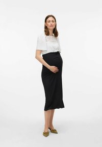 Outfit da maternità composto da una maglietta bianca testurizzata con dettagli in pizzo e una gonna nera aderente con arricciature laterali. Abbinato a ballerine verdi.