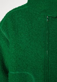 Veste polaire verte texturée avec une fermeture éclair argentée verticale et des détails visibles au niveau du col et des coutures.