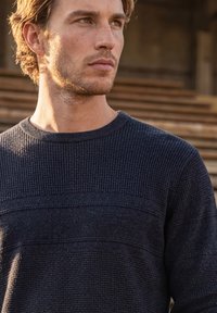 Jeune homme aux cheveux bruns courts portant un pull texturé bleu marine foncé, regardant sur le côté avec des escaliers en bois flous en arrière-plan.