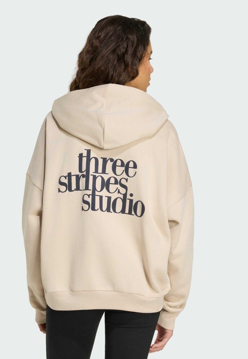 Krémově zbarvená volná mikina s kapucí a velkým černým nápisem „three stripes studio“ na zádech. Měkká textilie.