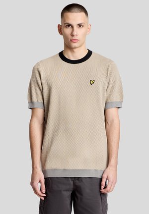 Giovane uomo con capelli corti che indossa una maglietta beige a maniche corte in maglia con colletto nero e piccolo logo di uccellino giallo, in piedi davanti a uno sfondo grigio.