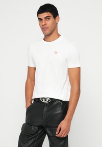 Biała bawełniana koszulka z krótkim rękawem, z małym czerwonym logo na lewej piersi. W połączeniu z czarnymi skórzanymi spodniami i srebrną sprzączką paska.