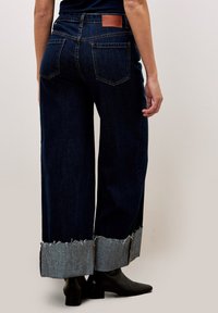 Dunkelblaue weit geschnittene Jeans mit fransigem Saum und kontrastierenden helleren Denim-Akzenten an den Knöcheln. Braunes Lederlogo-Patch auf der Rückseite.