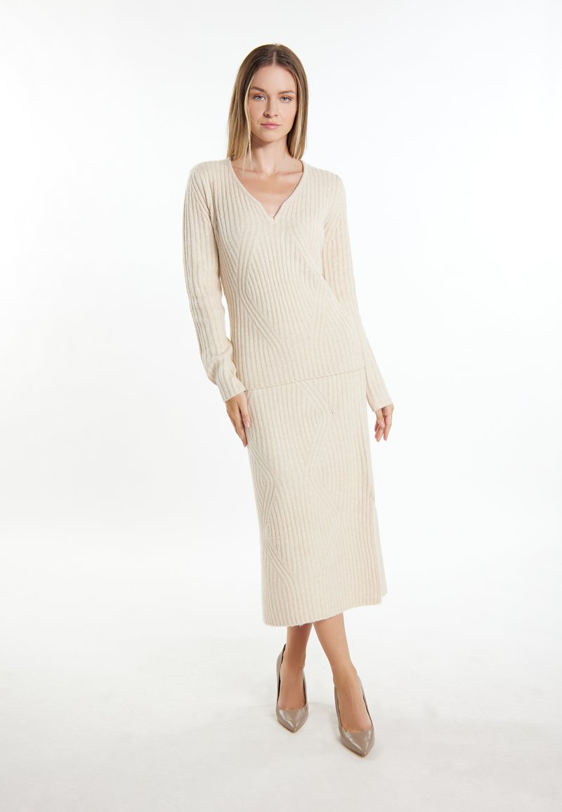 Strick-Zweiteiler in hellbeige mit gerippter Textur. Pullover mit V-Ausschnitt kombiniert mit einem Midirock, der ein Rautenmuster aufweist. High Heels in glänzendem Taupe.