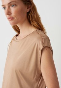 Lichtbruin top met korte mouwen, gemaakt van gladde stof, met een ronde hals en zachte plooien bij de schouders voor extra detail.