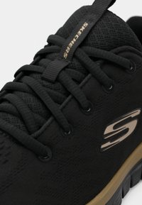 Baskets Skechers noires avec une tige texturée, des lacets plats noirs et un accent logo doré. La semelle en caoutchouc présente un bord beige contrastant.
