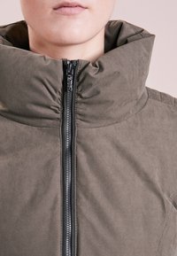 Närbild av en person som bär en helzipad höghalset taupe-färgad vadderad jacka mot en neutral bakgrund.