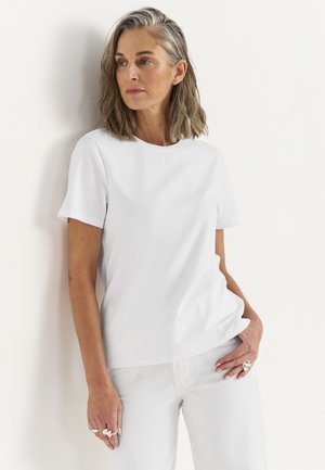 Kvinde med skulderlangt gråt hår iført en simpel hvid t-shirt og hvide bukser, stående op ad en hvid væg.