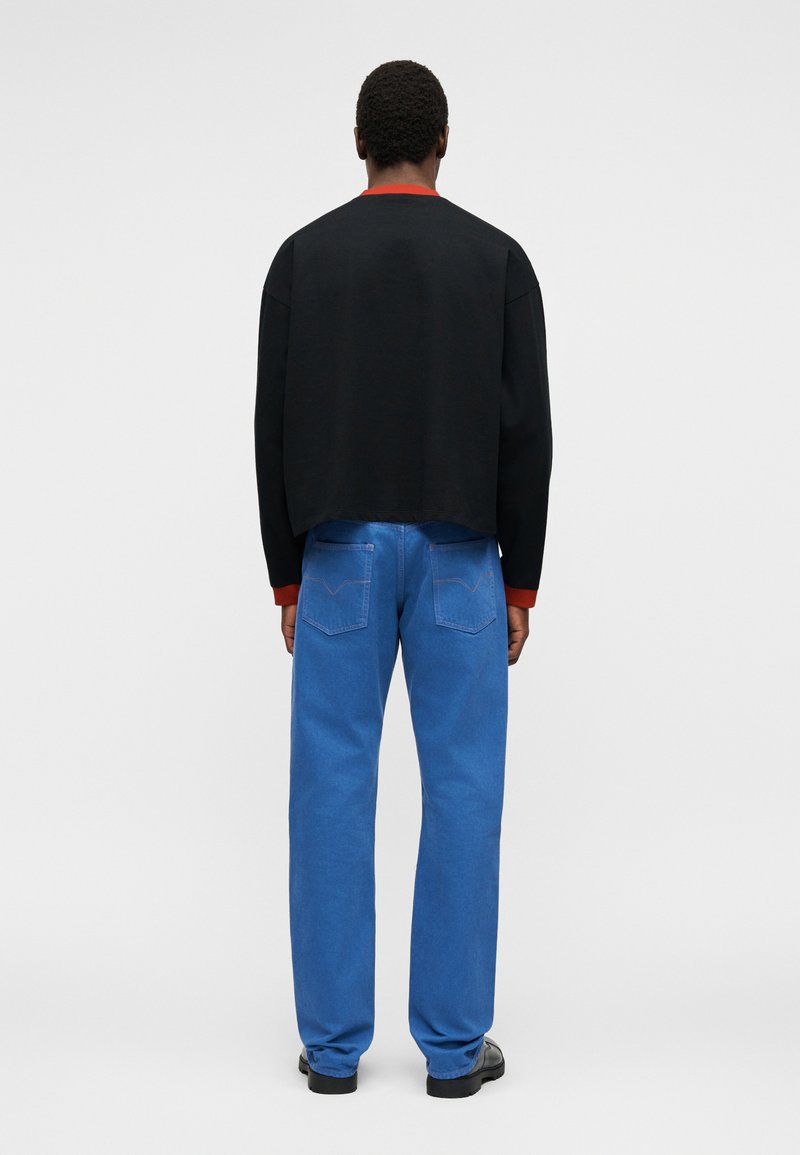 Homme debout de dos, portant une chemise noire à manches longues avec des poignets rouges et un jean bleu avec des poches arrière, chaussures noires, sur un fond clair.