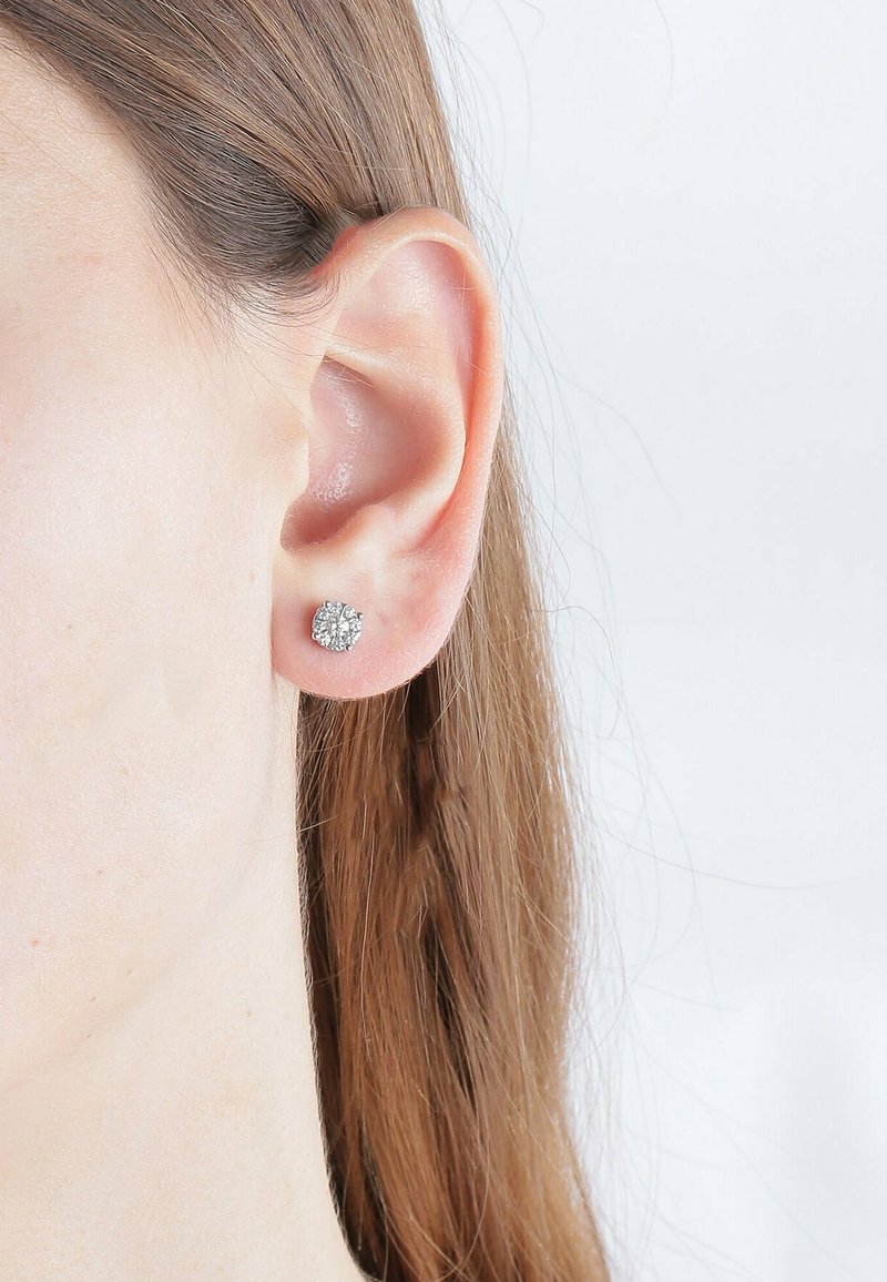 Linkes Ohr mit glatt gestrichenem, hellbraunem Haar, das hinter dem Ohr versteckt ist. Trägt einen kleinen runden silbernen Ohrstecker mit klaren Edelsteinen.