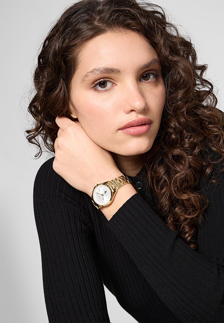 Reloj de oro con una esfera blanca redonda y marcas negras, que cuenta con una pulsera de metal enlazada. El modelo lleva una camiseta negra acanalada.