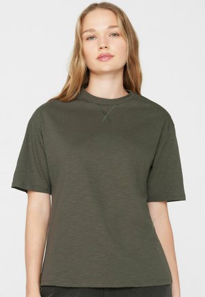 Vrouw met lang blond haar die een eenvoudige olijfgroene T-shirt met korte mouwen draagt, kijkt recht vooruit tegen een witte achtergrond.
