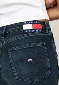 Los jeans de mezclilla oscura presentan un parche trasero con "TOMMY JEANS" en blanco y azul marino, junto con un logotipo rectangular en rojo, blanco y azul.