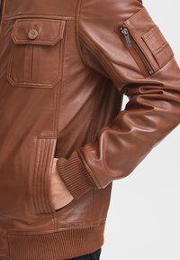 Hnedá kožená bunda s vreckom na zips na rukáve, rebrovanými manžetami a štruktúrovaným povrchom, ktorý zdôrazňuje elegantný, priliehavý strih.