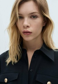 Svart skjorta med krage och guldknappdetaljer, utrustad med två framfickor. Tyget verkar slätt med en strukturerad design.