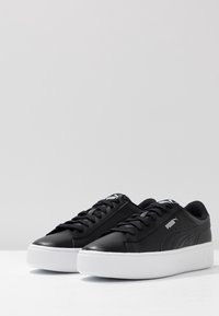 Sneaker in pelle nera con suole in gomma bianca, caratterizzati da una punta rotonda, design con lacci e un logo discreto sui lati.