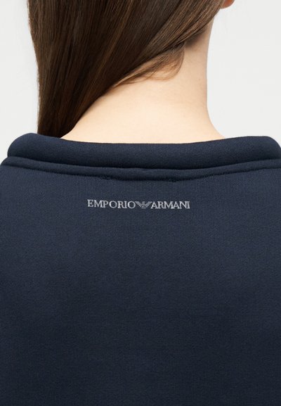 Πίσω όψη ατόμου με μακριά καστανά μαλλιά που φοράει μπλε σκούρο τοπ με το λογότυπο Emporio Armani στο πάνω μέρος της πλάτης.
