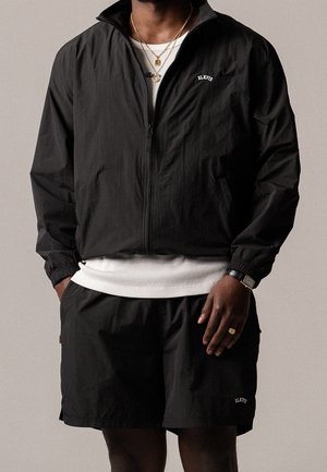 Man draagt een zwart zip-up jasje en bijpassende shorts over een wit hemd, gecombineerd met gouden kettingen, een ring en een polshorloge.