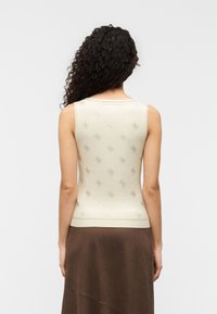Femme aux cheveux bruns bouclés, portant un haut beige sans manches avec un motif subtil et une jupe marron, vue de dos.