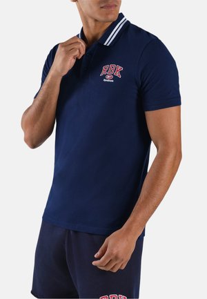 Uomo che sistema il colletto di una polo blu navy con strisce bianche e logo rosso "RBK Reebok", indossando pantaloncini blu navy coordinati.