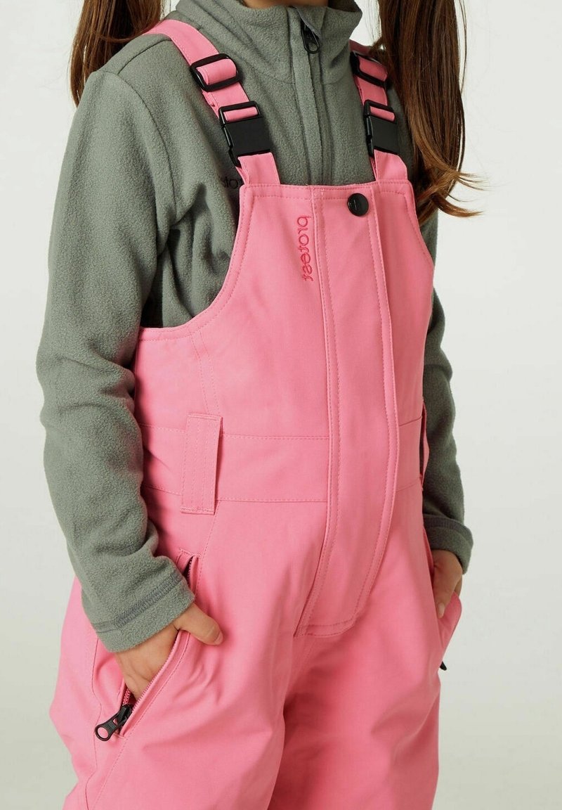 Niño con mono impermeable rosa con hebillas negras sobre una chaqueta de forro polar gris, con las manos en los bolsillos, de pie contra un fondo liso.