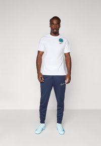 Hvid Nike t-shirt med blåt logo på brystet, parret med marineblå sweatpants og lyseblå træningssko. Minimalistisk design, ingen mønstre.