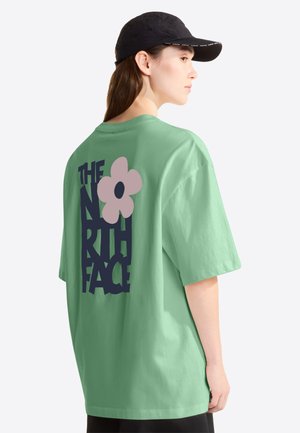 Žena nosí mentolově zelené tričko s nápisem "THE NORTH FACE" a růžovým květinovým motivem na zádech, doplněné černou čepicí.