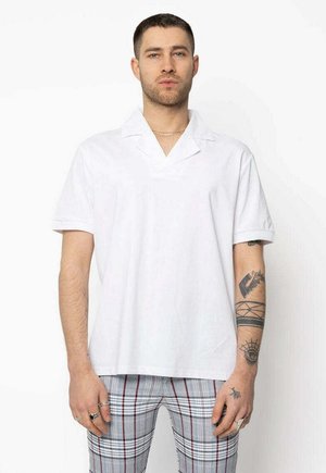 Un homme aux cheveux courts et tatoué, portant une chemise blanche à manches courtes et un pantalon à carreaux, se tient de face contre un fond blanc.