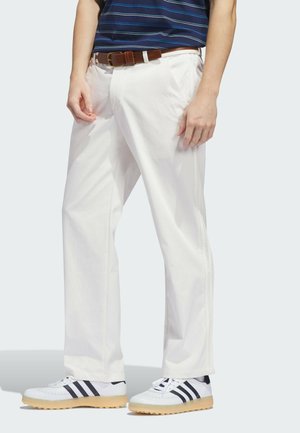 Uomo che indossa pantaloni bianchi a gamba dritta, cintura di pelle marrone e sneakers bianche con strisce nere, in piedi davanti a uno sfondo semplice.