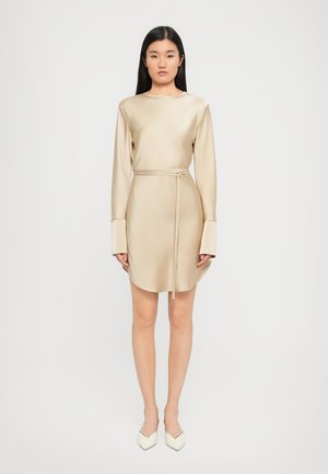 Robe en satin beige à manches longues et ceinture à la taille, avec des poignets larges et un ourlet légèrement arrondi. Portée avec des chaussures blanches à bout pointu.