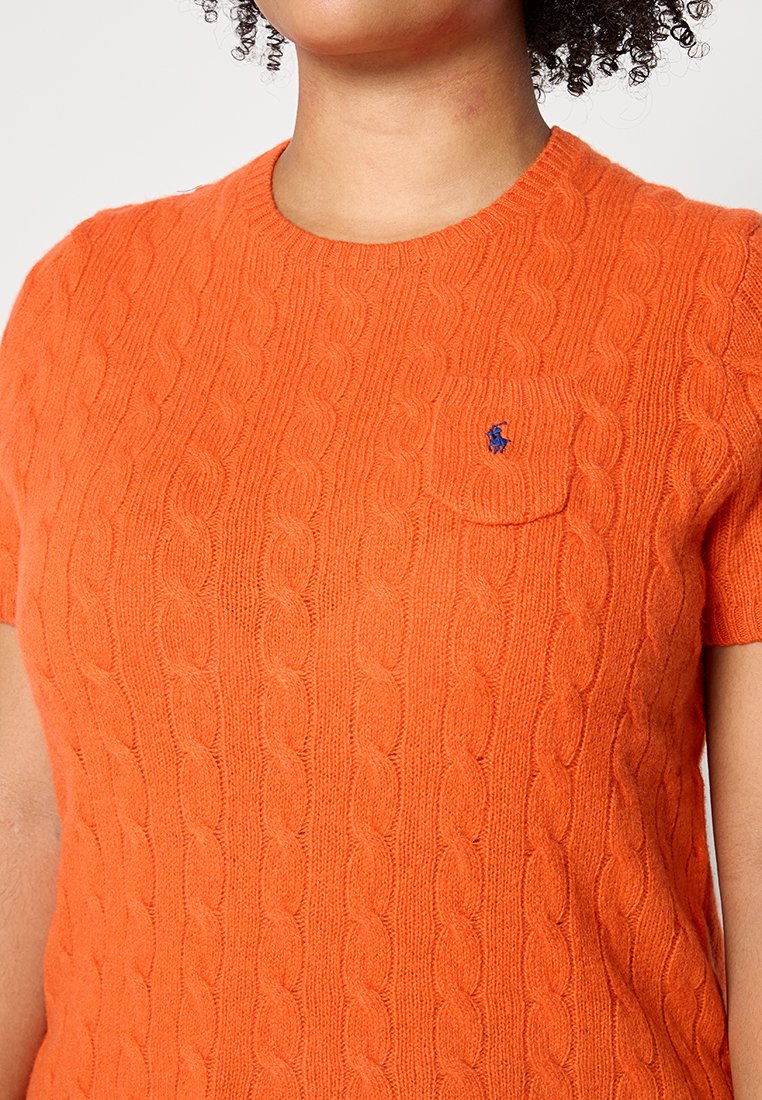Maglione arancione a maglia a maniche corte con motivo a treccia e scollo rotondo. Presenta una piccola tasca sul petto con un accento blu del logo.