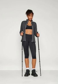 Svart idrettsjakke med glidelås, svart sportsbh, og svarte capri leggings. Modellen holder turstaver og har på seg svarte joggesko.