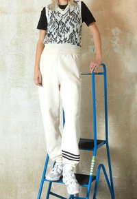 Pletená bezrukávová svetr s černobílým vlnkovým vzorem přes černé tričko; krémové tepláky s elastickým pasem; světle šedé tenisky.