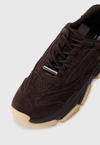 Zapatillas de ante marrón con paneles de malla, cordones redondos y un ojal metálico. Suela de goma de color claro con acentos texturizados.
