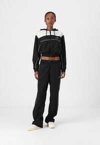 Cazadora negra con capucha y detalles en blanco, puños elásticos y diseño corto, combinada con pantalones negros con rayas laterales y cremalleras.