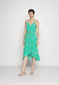 Vestido verde com estampa floral, alças finas, cintura com cinto e bainha assimétrica. Combinado com sandálias de salto bloco em rosa claro.